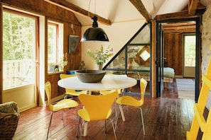 Interior de madera con una mesa redonda, sillas amarillas, una lámpara colgante negra grande y vistas a través de un ventanal.