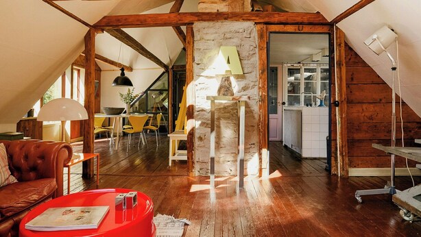 A loft-style room with exposed wooden beams, a stone wall, and a dining area with yellow chairs.