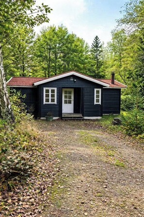 Eine kleine, dunkle Holzhütte mit rotem Dach, weißer Tür und Fenstern, umgeben von dichtem Wald.