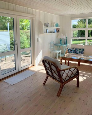 Ein Zimmer mit Holzboden, einem Korbsessel, einem Glastisch, einem blauen Sessel, einer Couch mit gemusterten Kissen und einem Regal mit Dekorationen.
