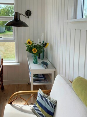 Ein Zimmer mit weißer Wand, einem Fenster, einem weißen Regal mit Büchern, einer Lampe und einer Vase mit Sonnenblumen.