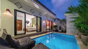 Un área moderna junto a la piscina con una piscina azul cristalina, un sofá y un espacio interior bien iluminado con una cama y decoración mural.