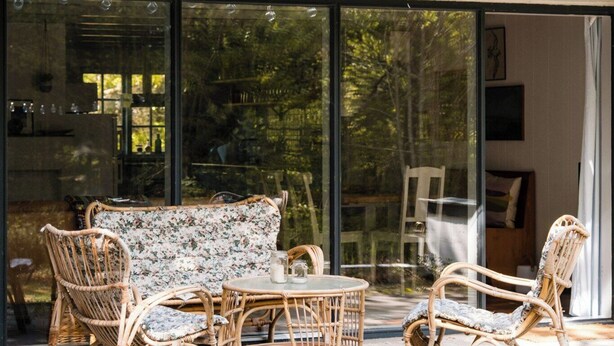 Un área de estar moderna al aire libre, con mobiliario de mimbre, una entrada con puerta de vidrio y un balcón con barandilla.