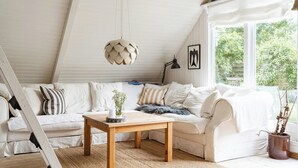 A white loft bedroom with a sloped ceiling, a wooden table, a white sofa, and a large window with a view of greenery. - Close to beach, forest and bike trails (Grenå)