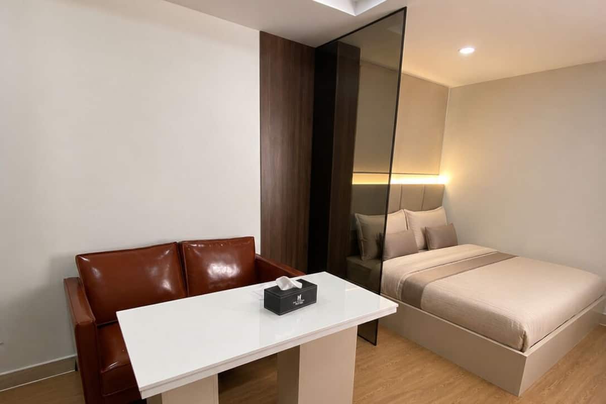 A modern room with a brown leather couch, a white table with a tissue box, a bed with beige bedding, and a wooden floor.