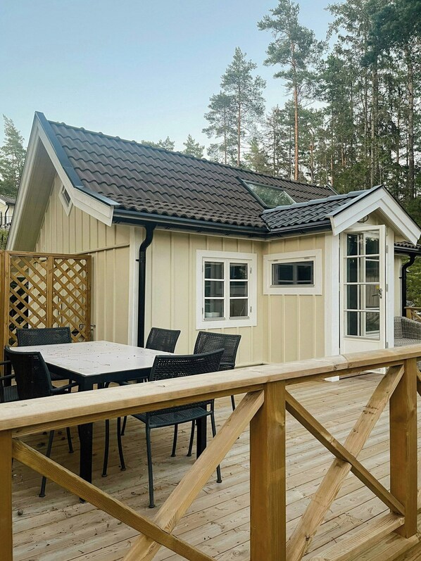 A wooden deck with a table and chairs, a small building with a sloped roof, and a wooden fence. - Cabin with private beach at Ingarö (Ingar ö)