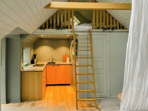 A loft bedroom with a wooden ladder, a bunk bed, a kitchen area, and a door. - Cabin with private beach at Ingarö (Ingarö)