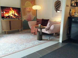 Ein Wohnzimmer mit Fernseher, der ein Feuer zeigt, einer Couch, einem kleinen Tisch mit einer Tasse, einem gemusterten Teppich und einer Kiefernzapfen-Dekoration.