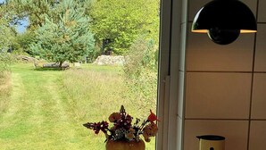 A kitchen with a window view of a grassy yard and trees, a vase with flowers, a bottle, and a mug on the counter. - Retro guest house near Rebild Hills (Hadsund)