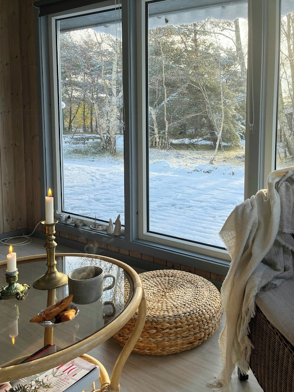 Ein gemütliches Zimmer mit großem Fenster, einem Glastisch mit Kerzen und einem gewebten Pouf.