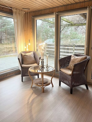 Ein Zimmer mit Korbmöbeln, einem Glastisch und Blick auf eine Holzterrasse und Bäume.