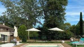 A rectangular swimming pool with a wooden deck, two white umbrellas, and a fenced area with greenery and a building in the background. - Unique gite in a renovated factory in Provence (Salernes)