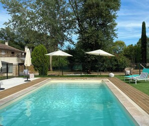 A rectangular swimming pool with a wooden deck, two white umbrellas, and a fenced area with greenery and a building in the background. - Unique gite in a renovated factory in Provence (Salernes)