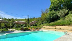 Piscina trasparente con lettini e ombrelloni, immersa nel verde.