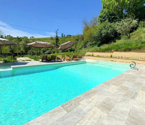 A clear swimming pool with lounge chairs and umbrellas, surrounded by greenery.