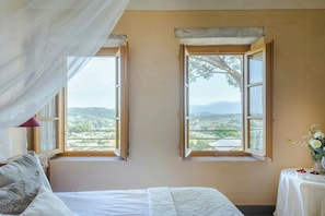 Ein Schlafzimmer mit einem Bett, zwei Fenstern, einem Nachttisch mit einer Vase und Blick ins Freie.