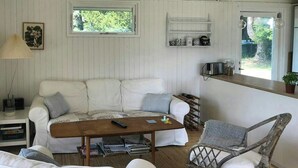 Et hyggeligt rum med en hvid sofa, et træbord, en fletstol og en lille hylde med pynteting.