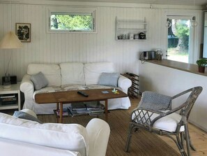 Ein gemütliches Zimmer mit einer weißen Couch, einem Holztisch, einem Korbstuhl und einem kleinen Regal mit Dekoration.