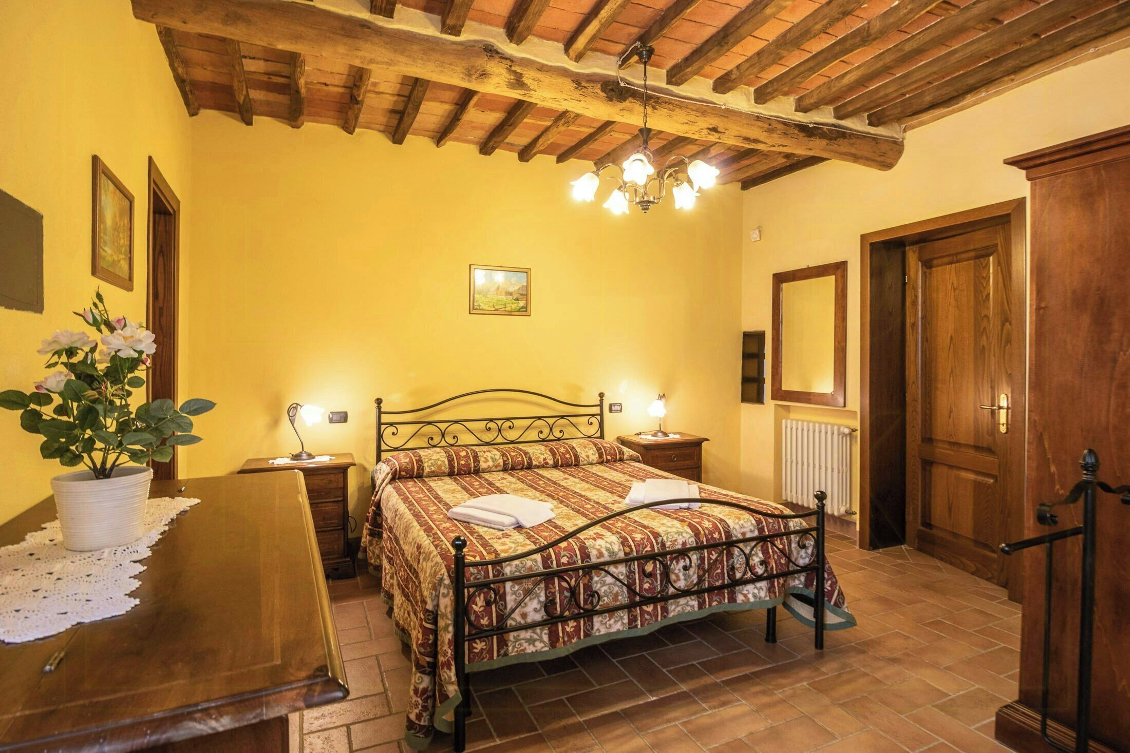 A bedroom with a wooden ceiling, a bed with a patterned bedspread, a bedside table with a lamp, and a potted plant.