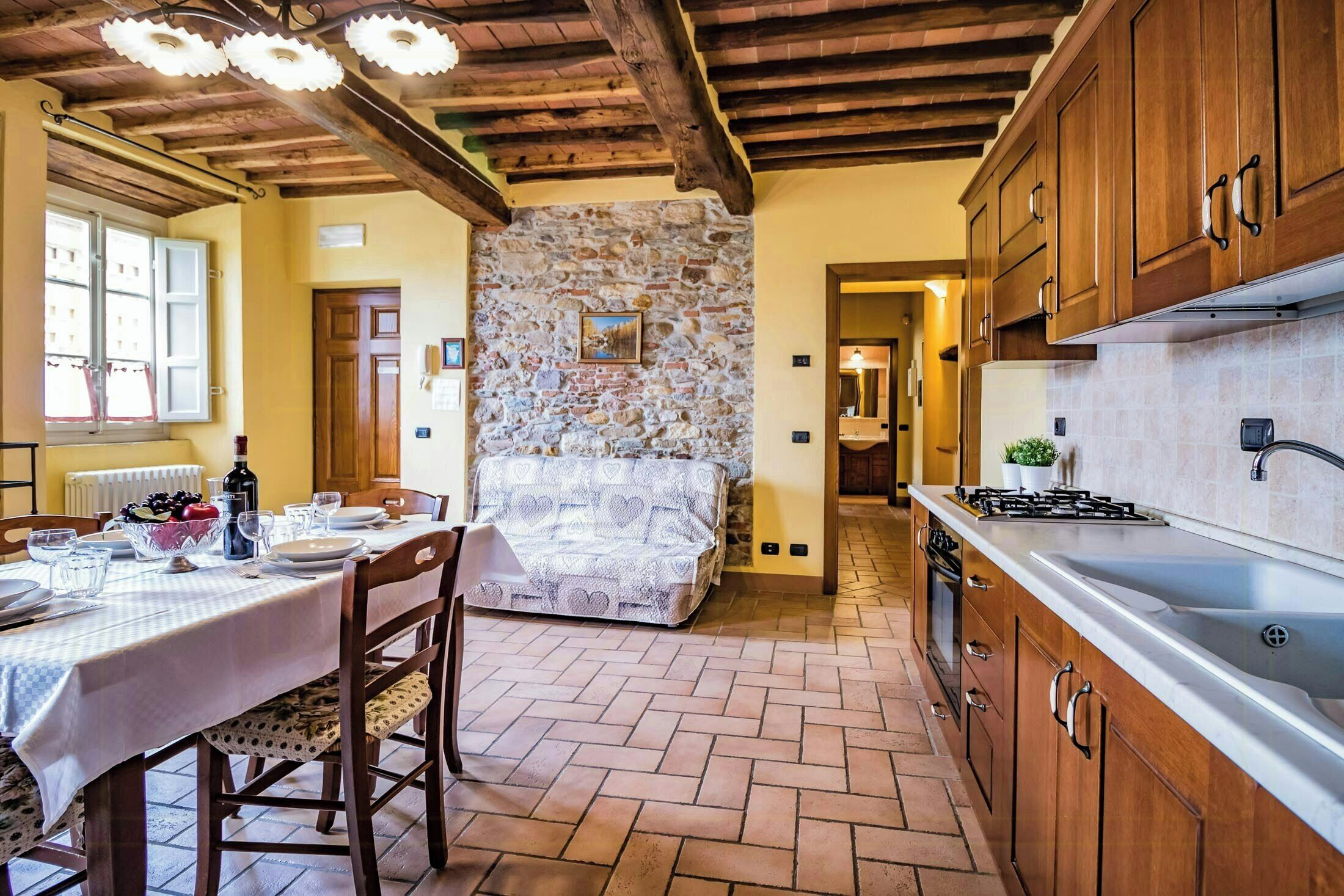 A rustic kitchen with wooden cabinets, a stone wall, and a tiled floor.