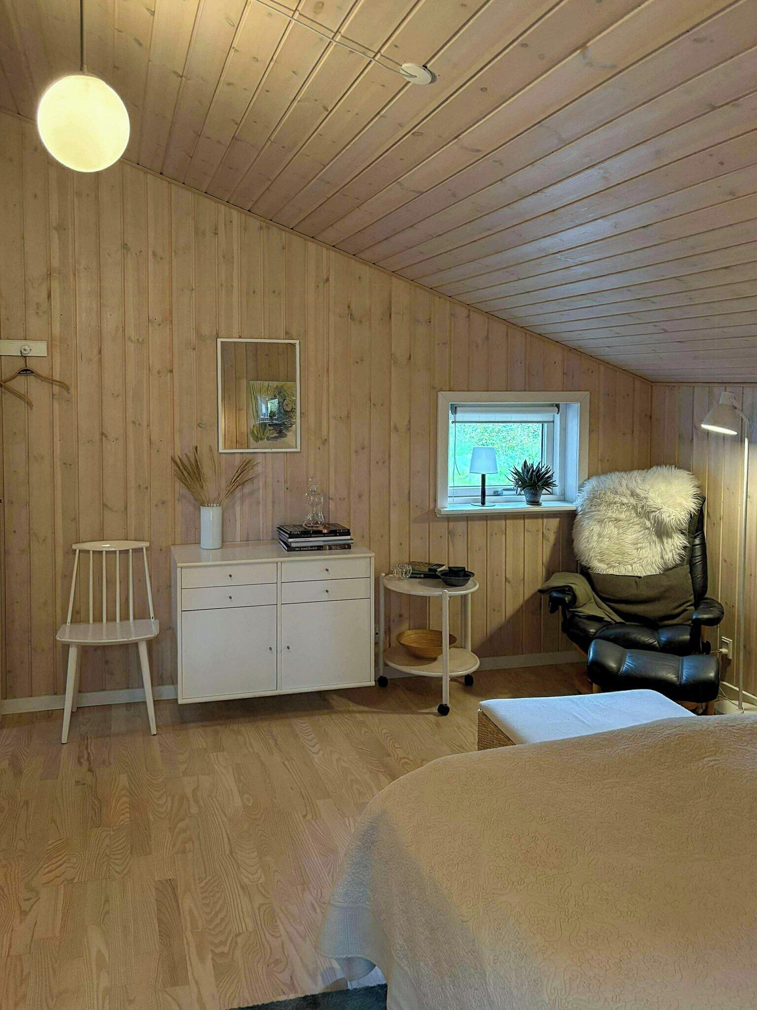 Et værelse med trævægge og -loft, et hvidt skab, en stol, et lille bord og et vindue med udsigt over grøn vegetation.