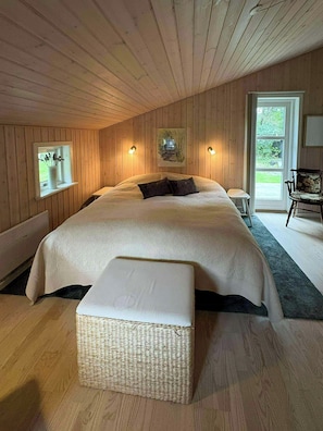 Ein Schlafzimmer mit einem großen Bett, einem kleinen Nachttisch, einem Sessel und einem Fenster mit Blick auf Grünfläche.