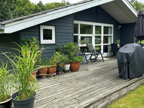 Eine Holzterrasse mit Blumentöpfen, einem überdachten Grill und Gartenmöbeln.
