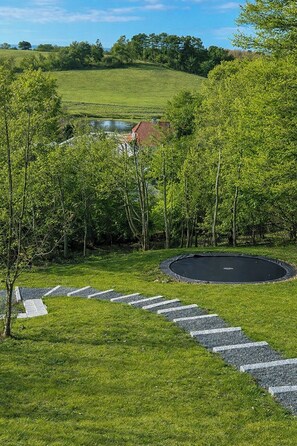 Eine Rasenfläche mit Trampolin, Teich und einem Gebäude im Hintergrund mit rotem Dach.