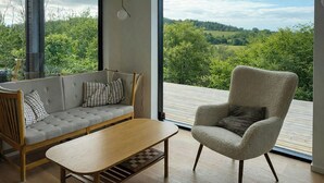 Ett modernt rum med en soffa, fåtölj och ett träbord, stora fönster med utsikt över grönområden.