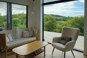 Ein modernes Zimmer mit einer Couch, einem Sessel und einem Holztisch, große Fenster mit Blick auf Grünflächen.