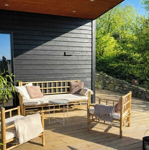 Eine Holzterrasse mit einer Couch und Stühlen, eine Steinmauer und ein Gebäude mit dunkler Holzfassade.