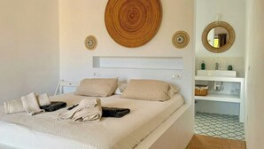 Un llit ben arropat amb tovalloles doblegades, una decoració circular de fibra natural i un mirall sobre una prestatgeria blanca amb productes de tocador.