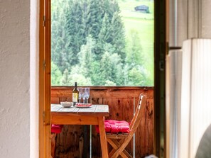 Une table en bois avec une bouteille, des verres et une assiette de nourriture, un coussin rouge et une chaise, avec vue sur une forêt par la fenêtre.