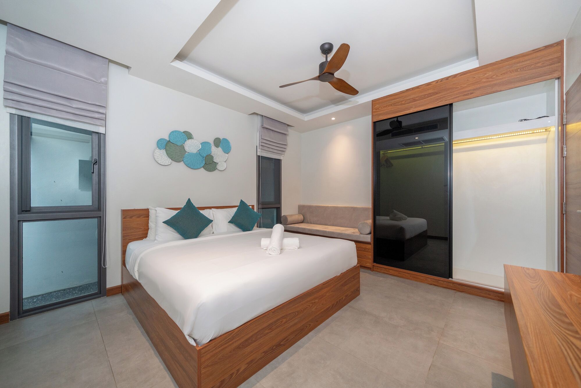 Une chambre moderne avec un lit en bois, un ventilateur de plafond, une œuvre d’art accrochée au mur et un placard à portes coulissantes.