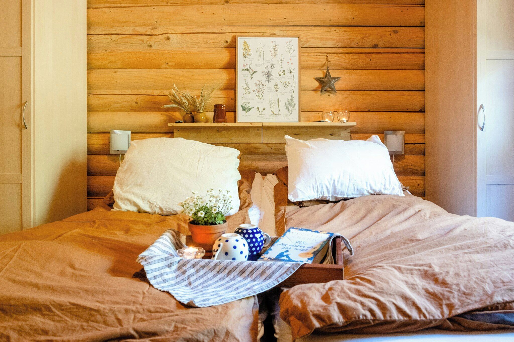 A wooden-paneled room with a bed, pillows, a framed botanical illustration, a star decoration, and a bedside table with a plant and a candle.