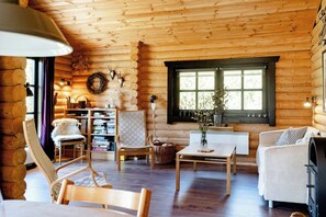 A log cabin interior with a fireplace, a bookshelf, a dining table, and a cozy seating area. - Authentic log house amid evergreens in Vendsyssel (Fjerritslev)