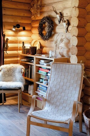 A cozy cabin interior with a wooden wall, a woven chair, a bookshelf, and a fireplace. - Authentic log house amid evergreens in Vendsyssel (Fjerritslev)