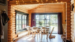 Een interieur van een blokhut met een eethoek, houten wanden, grote ramen en een kroonluchter.