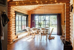 A log cabin interior with a dining area, wooden walls, large windows, and a chandelier. - Authentic log house amid evergreens in Vendsyssel (Fjerritslev)