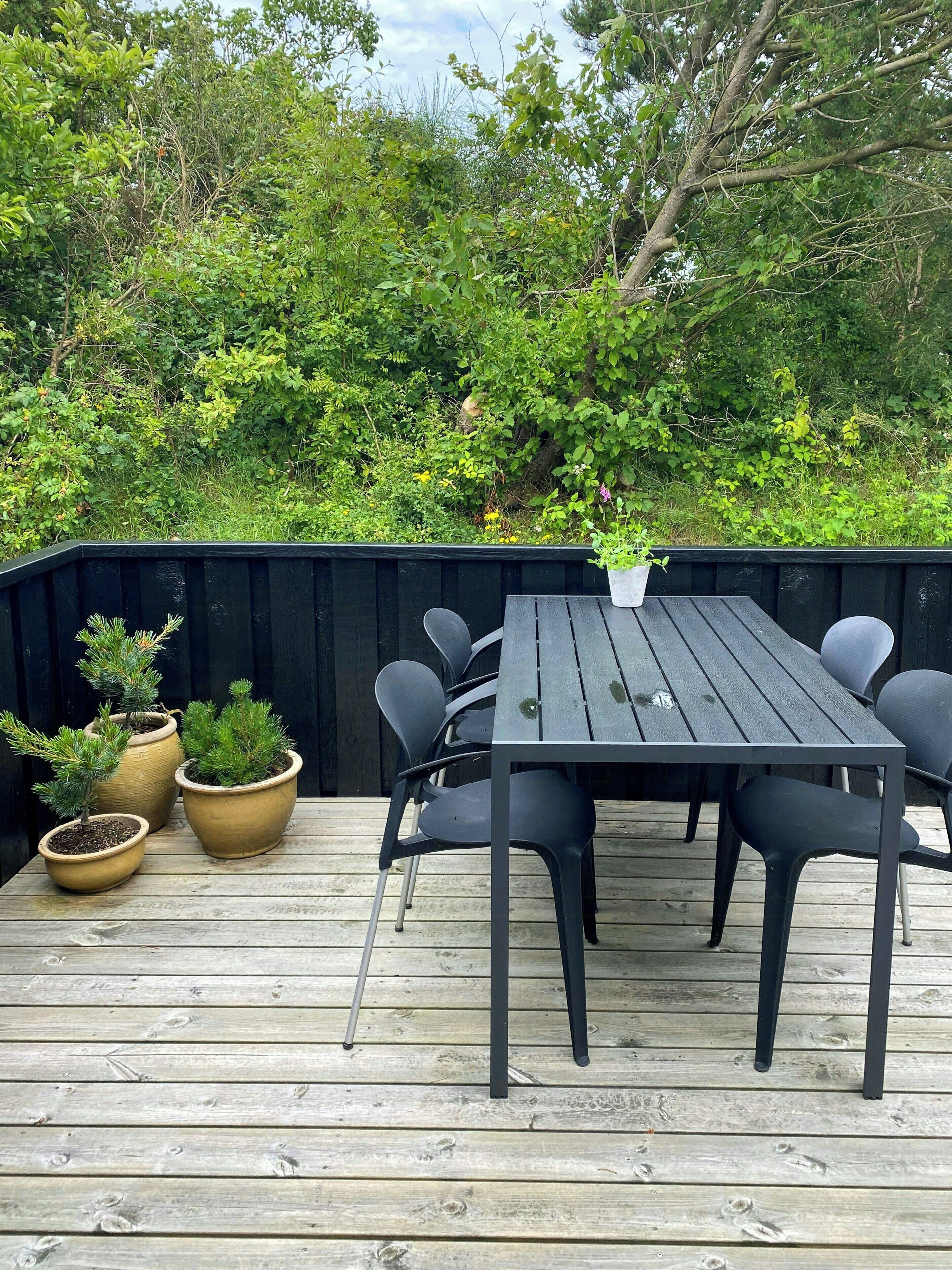 A wooden deck with a table and chairs, surrounded by greenery and a black fence.