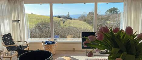 A room with a view of a green landscape, a black bowl, a wicker basket, a chair, a table, and a vase with flowers.