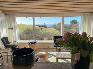 Ein Zimmer mit Blick auf eine grüne Landschaft, eine schwarze Schüssel, ein Korbwägelchen, ein Stuhl, ein Tisch und eine Vase mit Blumen.