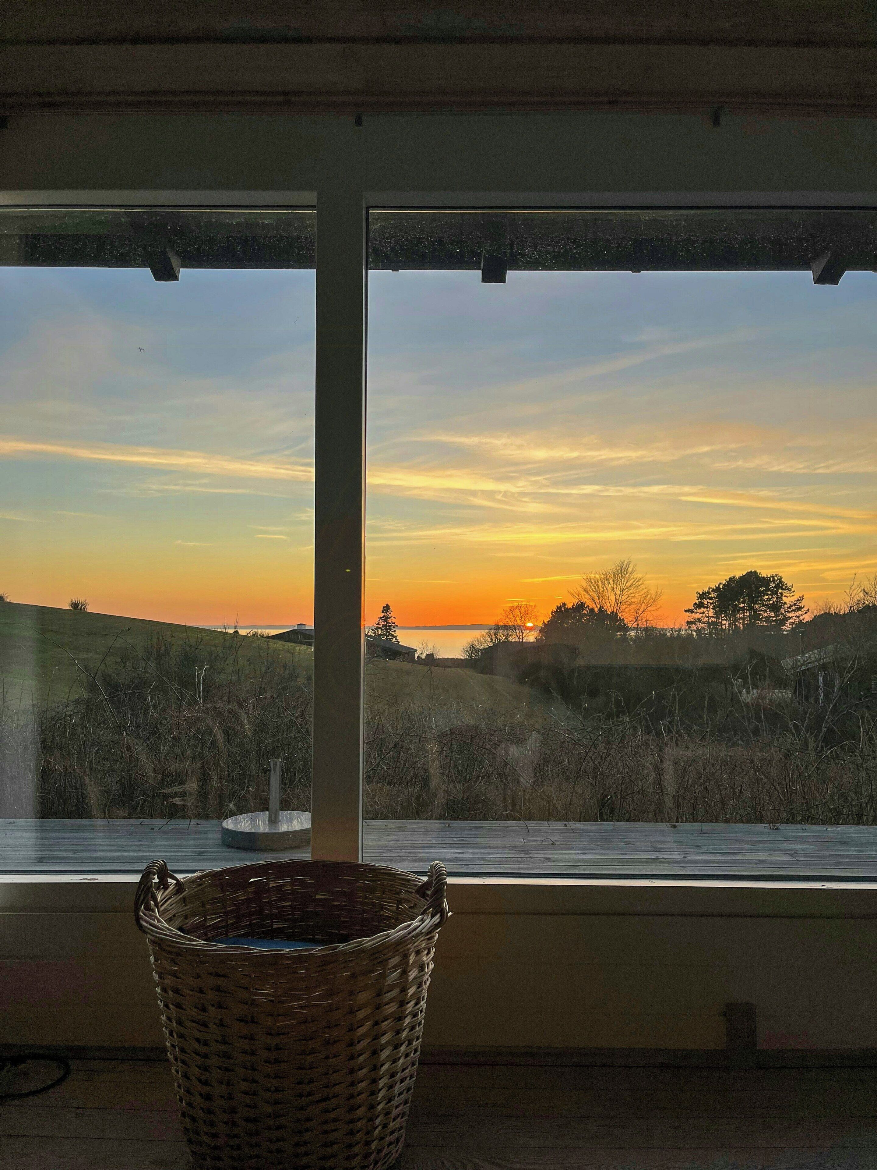 Une pièce dotée d’une grande fenêtre donnant sur un paysage au coucher du soleil, un panier en osier posé sur le rebord de la fenêtre et une cheminée visible.