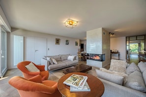 Ein modernes Wohnzimmer mit Sofa, Sessel, Couchtisch, Kamin und einem Essbereich mit Stühlen.