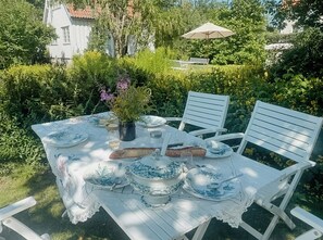 Een buitentafel gedekt met witte stoelen, omringd door groen en een wit gebouw op de achtergrond.
