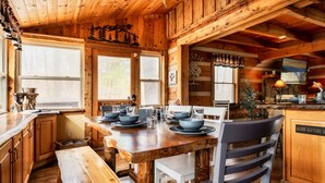Ein Holzblockhaus-Interieur mit einem Esstisch, gedeckt für vier Personen, Holzwänden und einer Küchenzeile. Ein Schild trägt die Aufschrift 'WELCOME TO'.