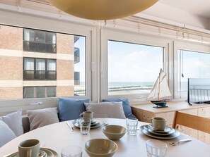 A dining area with a view of the sea, a sailboat model, and a television on a wooden table. - Sunny corner apartment with luxury sea view. (Blankenberge)