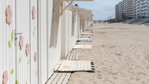 Eine Reihe von Strandhäusern mit Holzstegen, die zu ihnen führen, vor einem Sandstrand und einem großen Gebäude im Hintergrund.
