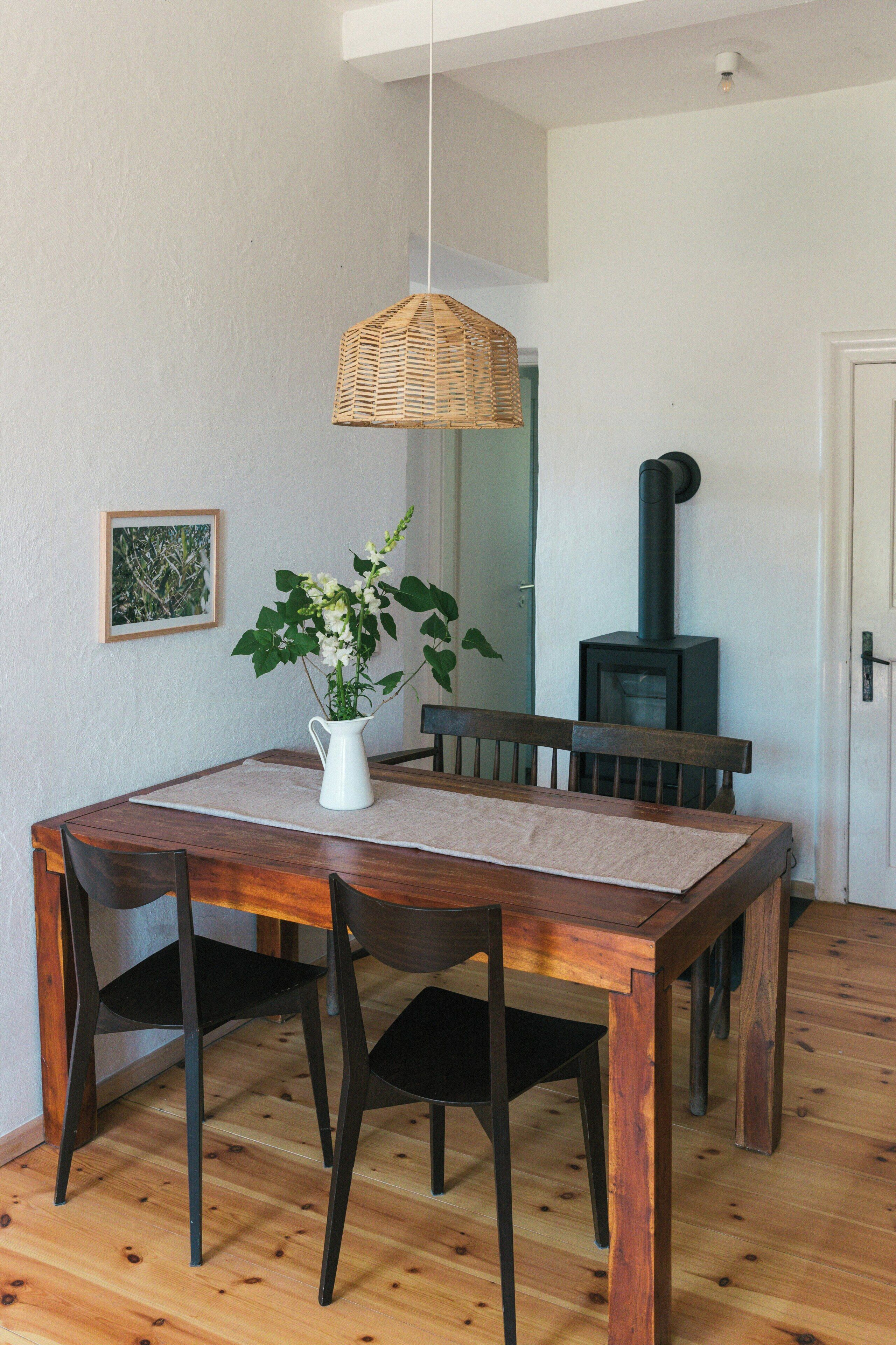 Een eetruimte met een houten tafel, stoelen, een hanglamp, een vaas met bloemen, een lijst met een afbeelding en een houtkachel.