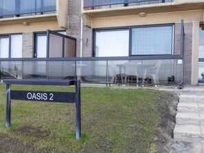 Ein modernes Gebäude mit einem Schild, auf dem 'OASIS 2' steht, großen Fenstern und einer Rasenfläche mit Gartenmöbeln.